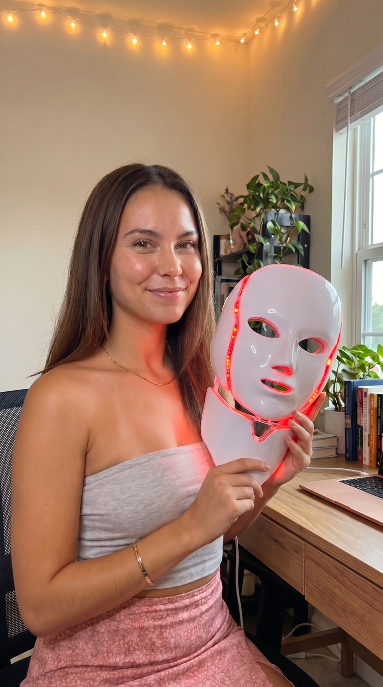 Woman holding the glowing NeoTelle LED light therapy mask in a warm home setting