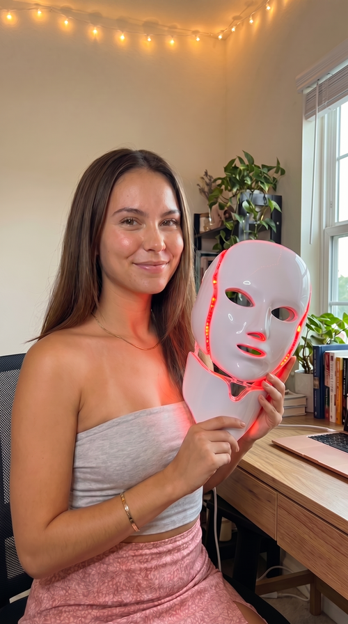 Woman holding the glowing NeoTelle LED light therapy mask in a warm home setting